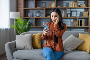 Surprised asian woman using smartphone at home. Sitting on sofa, reading unexpected message on...