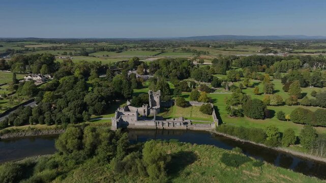 Desmond Castle, Adare, County Limerick, Ireland, September 2024. Drone orbits clockwise in a medium establishing view of the ruins on the River Maigue with water reflecting trees under clear blue sky.