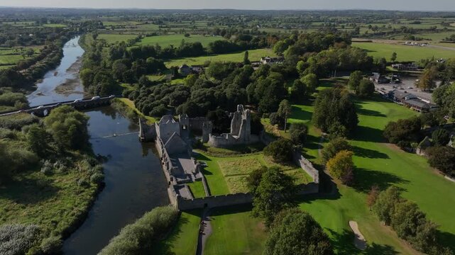 Desmond Castle, Adare, County Limerick, Ireland, September 2024. Drone orbits clockwise and pulls back showcasing the historic ruins on the edge of River Maigue under a clear blue sky.