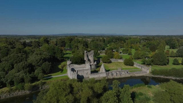 Desmond Castle, Adare, County Limerick, Ireland, September 2024. Drone orbits clockwise and descends over trees by the River Maigue with the white grey ruined stone walls along the riverbank.