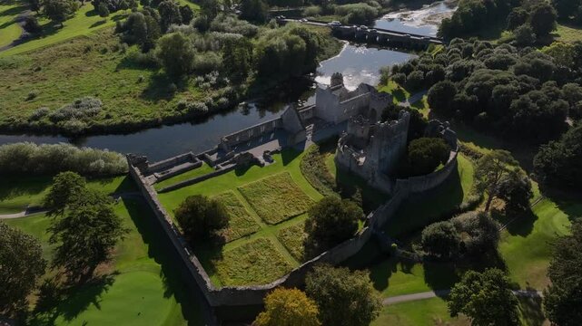 Desmond Castle, Adare, County Limerick, Ireland, September 2024. Drone orbits clockwise in a high angle view of ruins with imposing stone walls on the River Maigue as sunlight sparkles on the water.
