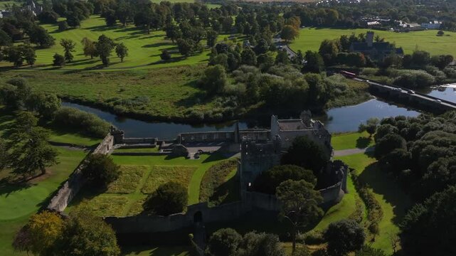 Desmond Castle, Adare, County Limerick, Ireland, September 2024. Drone orbits clockwise in a high angle of the ruins by the River Maigue as sunlight shines on the water with forest and grassy fields.