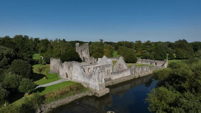 Desmond Castle, Adare, County Limerick, Ireland, September 2024. Drone orbits clockwise and descends passing the trees by the River Maigue to establish the crumbling stone walls under a clear blue sky