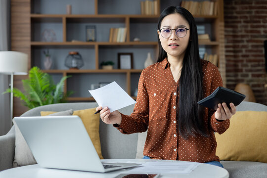 Asian woman at home looks puzzled over finances, holding paper and calculator. Laptop on table representing remote work challenges and financial planning stress.