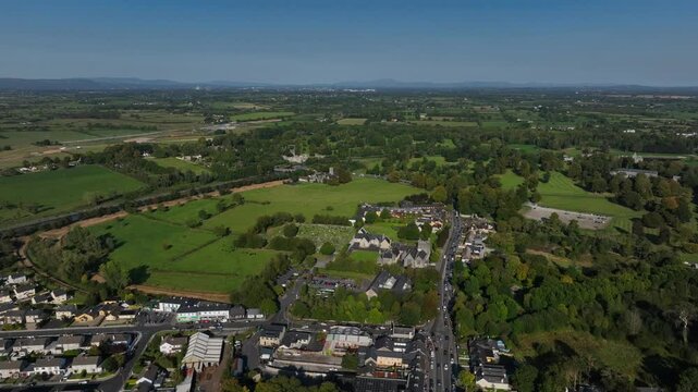 Adare, County Limerick, Ireland, September 2024. Drone pushes forward to the north west above the village moving over a cemetery and open fields along the main street with scattered woods and farms.