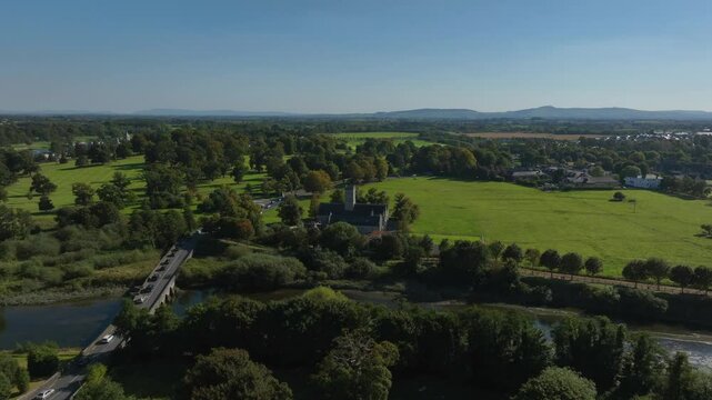 Augustinian Friary, Adare, County Limerick, Ireland, September 2024. Drone orbits clockwise crossing the stone bridge as traffic exits the village over the River Maigue winding through the countryside