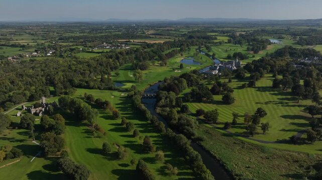 Adare, County Limerick, Ireland, September 2024. Drone orbits counter clockwise pulling back above friary ruins and manor beside the river with line of trees casting shadows on the golf course.