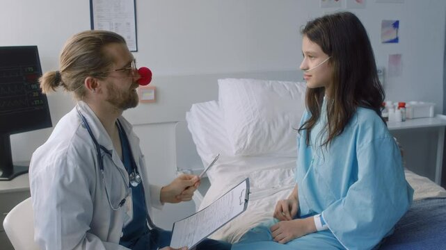 Side shot of cheerful male pediatric neurologist with red nose checking attention focusing of little smiling girl, doctor touching her nose with pen and she squeezing doctors red nose inclinic