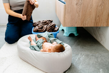 Mother doing laundry while baby relaxes on cozy pillow.