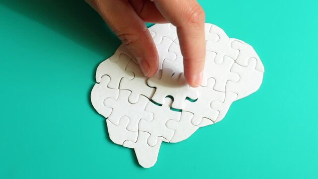Close-up of a hand placing a missing puzzle piece into a brain shape on a teal background, illustrating cognitive engagement and problem-solving.