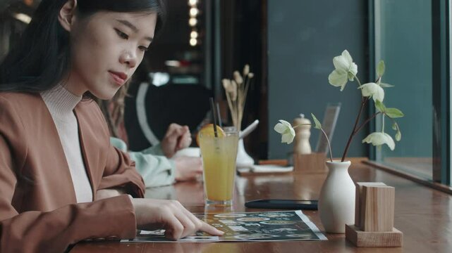 Young Asian woman sitting at table with cocktail in busy restaurant and looking at images of dishes on menu, choosing what else to order