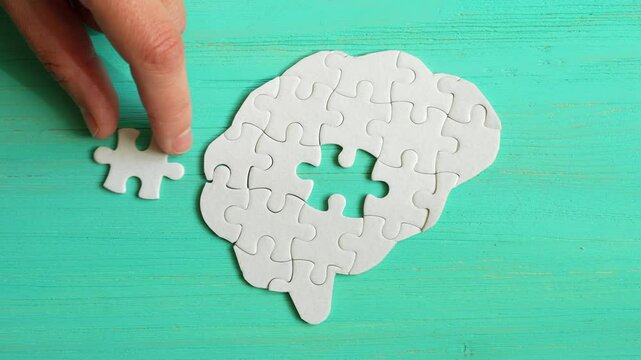 Close-up of a hand taking the final jigsaw puzzle piece to insert into a brain-shaped puzzle on a teal background to complete it.