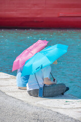
two ladies sit with pink and blue umbrellas on the edge of the harbor quay