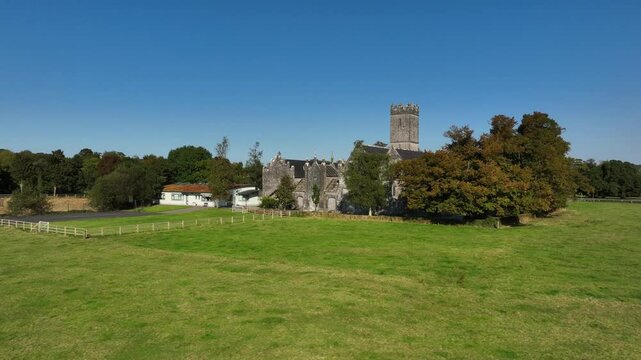 Augustinian Friary, Adare, County Limerick, Ireland, September 2024. Drone orbits clockwise at a low level showcasing the grand gothic stone church from behind trees under a perfect blue sky.