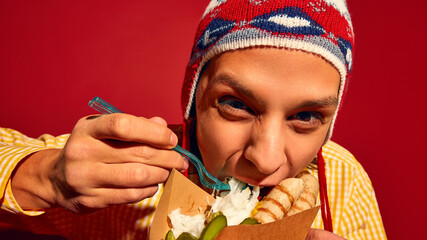 Close-up of emotive young man in winter hat and yellow sweater eating passionately sausages, Bratwurst with pickles and sauerkraut against red background. Concept of street food, winter, pop art