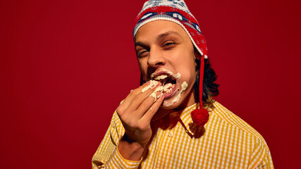 Delicious messy bite. Young man in winter hat, with face covered in mayonnaise sauce, eating...