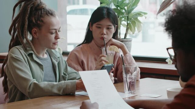 Man sitting at table and looking through restaurant menu, while his female friends talking to each other, sipping cocktails and looking at smartphone