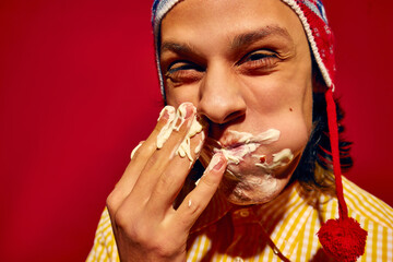 Delicious messy bite. Young man in winter hat, with face covered in mayonnaise sauce, eating delicious sausage, Bratwurst against bright yellow background. Concept of street food, winter, pop art