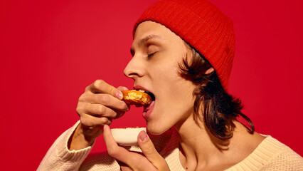Young guy in red beanie hat enjoying donuts dipped in sweet sauce, set against bold red background. Ideal for street food and holiday market promotions. Concept of street food, winter, pop art