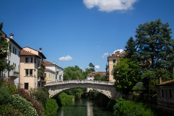 A small bridge and a river next to a street and facades of houses in the Italian city of Vicenza in the Veneto region in summer against a background of blue sky and green trees.