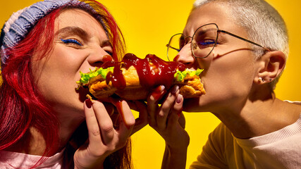 Super delicious street food. Two women emotional and passionately eating hot dog, biting it from...