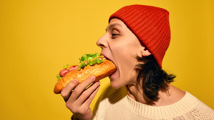 Young man in red beanie hat emotionally and passionately eating hot dog, set against bright yellow background. Concept of street food, winter, pop art, fairs, emotions, Christmas. Copy space