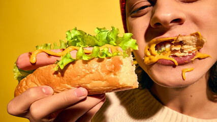 Young man in red beanie hat eating hot dog with mustard and lettuce, set against bright yellow background. Close-up. Concept of street food, winter fairs, pop art, holiday snacks