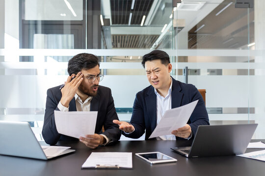 Businessmen in office discuss report, showing frustration over business performance. Laptops and phone on table emphasize work environment. - Powered by Adobe