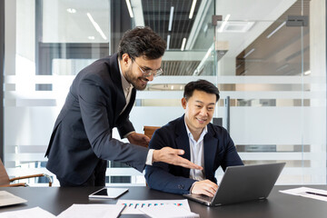 Businessmen collaborating in office. Focused team discussing project at laptop. One standing, one sitting, with smartphone and documents on table. Professional teamwork concept.