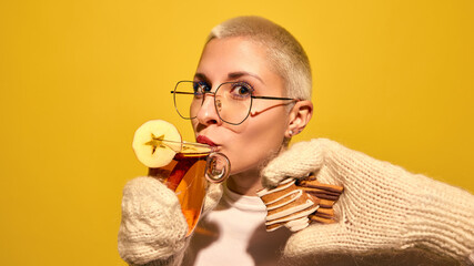 Young woman with short hair, in knitted gloves sipping hot cider and holding gingerbread cookies against bright yellow background. Concept of street food, winter fairs, pop art, holiday snacks