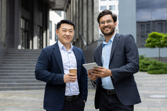 Two businessmen having an outdoor meeting, one holding coffee, other with digital tablet. Both smiling in business attire, representing teamwork, cooperation, and professional interaction.