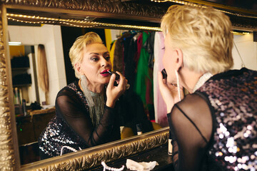 In a bustling backstage setting, a drag queen expertly applies bright lipstick while prepping.