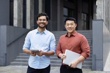 Confident business professionals standing outdoors with digital devices, smiling in modern urban setting. Men showcasing teamwork and collaboration in business environment.