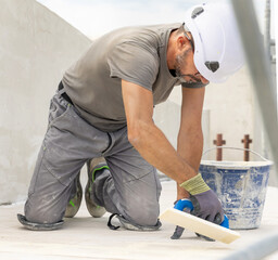 Craftsman in grey work clothes puts grout with a float sitting on his knees during construction