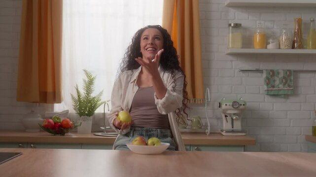 Hispanic Caucasian happy smiling funny woman girl female holding healthy food fruits at kitchen home joy throw juggle juggling apples throwing weight control diet delivery nutrition health cooking
