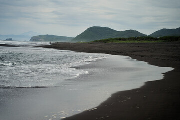 Khalaktyrsky Beach, Kamchatka. A tranquil coastal scene showcases black sand, gentle waves, and beautiful lush green hills set in the backdrop