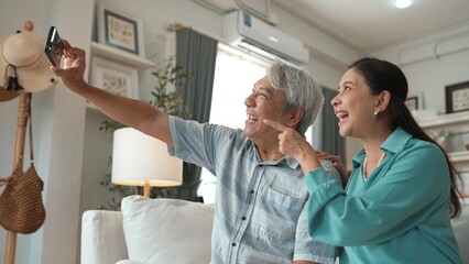 Obraz premium Happy grandfather and grandmother making video call with their son at home. Elder man holding smart phone and sitting at sofa while old woman greeting with cheerful vibe at modern house. Myrmidon.