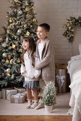 Little boy and girl hugging standing near the Christmas tree in a cozy modern interior.