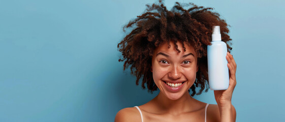 A woman with curly hair holds a white bottle of shampoo, smiling and covering half her face on one side against a blue background.