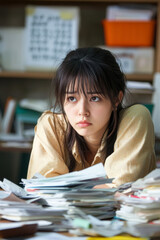 A Japanese young teacher woman sitting on her desk, her shoulders slumped as she stares at a messy pile of papers, with an unorganized classroom visible in the background