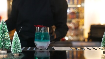 Bartender serving Christmas blue Lagoon cocktail with red dried fruit rosemary design on luxury bar counter with VIP group guest friend or couple in green neon snow Christmas tree festive. Vinosity.