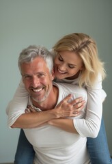 A cheerful older man beams with joy as a younger woman playfully hugs him from behind, their smiles radiating warmth and happiness. The soft, muted background enhances the intimate atmosphere