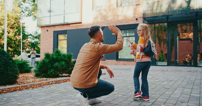 Dad saying goodbye to his daughter in schoolyard. Happy little girl waving hand to dad and running to school to study. Schoolgirl with backpack holding teddy bear with both hands. Studying day.
