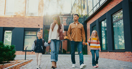 Front camera view of parents taking their kids from school. Caucasian man talking with woman while walking together in same direction. Children happy finishing difficult study day.