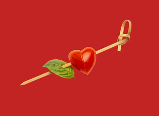 Heart shaped tomato and a fresh basil leaf are skewered together on red background, creating a simple elegant appetizer, a romantic valentine's day celebration