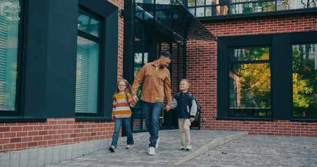 Front camera view of handsome Caucasian father walking away from school together with his charming children. Everyone smiling. Going back home after studying. Warm autumn weather. Carrying backpacks.