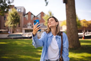 Fototapeta premium Image of smiling young blonde hair woman taking selfie photo holding her mobile phone while walking in park. Girl make blow kiss, air kiss, look happy.