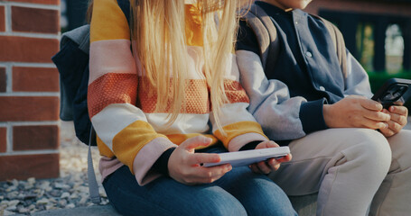 Side camera view of two Caucasian kids waiting for someone after school. Looking at smartphones. Holding mobile devices horizontally with both hands. Girl with glasses glancing directly.