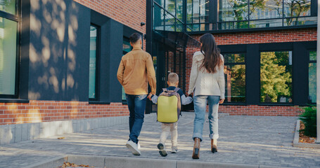 Young family spending time together while walking outdoors near contemporary building with red brick walls and black window frames. Child going between parents. Ready for school with his backpack.