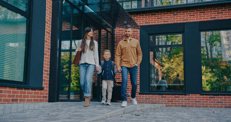 Family of three people walking outside next to building with large windows. Pretty mother in white...